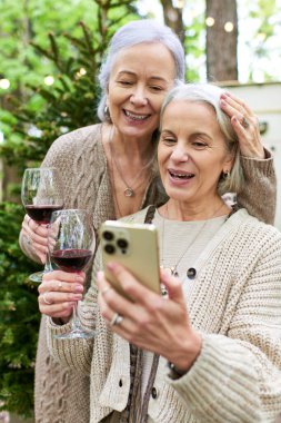 Two middle-aged women, a lesbian couple, relax and enjoy wine and technology while camping in a lush green forest. clipart