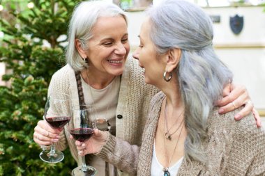 Two middle-aged women laugh together while enjoying wine outdoors near a camping van in a green forest. clipart