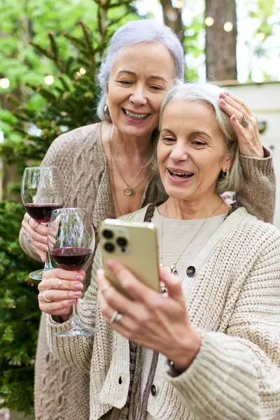 stock image Two middle-aged women, a lesbian couple, relax and enjoy wine and technology while camping in a lush green forest.