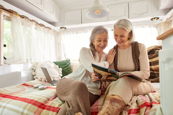 Две женщины среднего возраста, лесбийская пара, счастливо отдыхают в своем фургоне во время поездки. Они сидят на кровати и читают журнал..