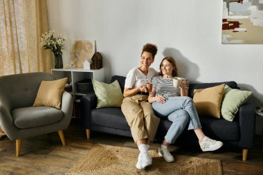 A diverse lesbian couple enjoys a relaxing afternoon at home, sharing coffee and smiles on the couch. clipart