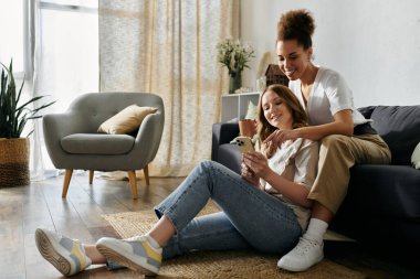 İki kadın evde dinleniyor, biri yerde oturuyor ve diğeri de bir kanepede oturup birlikte telefona bakıyor..