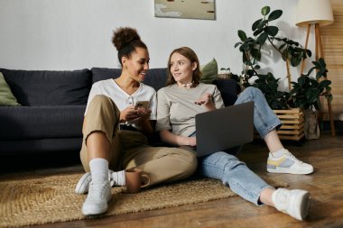 A lesbian couple relaxes at home, one using a laptop, the other on their phone. clipart