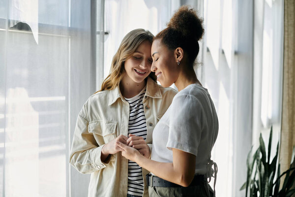 Two women in casual clothing stand close to each other by a window, smiling and holding hands.