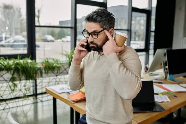 Sakallı yakışıklı bir işadamı elinde kahve fincanıyla modern bir ofiste dikilirken telefonuyla konuşuyor..