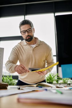Sakallı bir işadamı modern ofis ortamında dururken belgeleri dikkatle inceliyor..