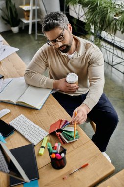 Sakallı yakışıklı bir işadamı modern bir ofiste çalışıyor. Elinde bir fincan kahve tutarken renk paletinden renkler seçiyor..