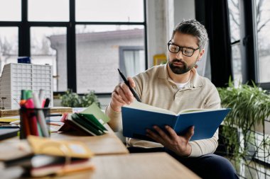 Sakallı yakışıklı bir işadamı modern bir ofiste masa başında oturmuş kitap okuyor ve düşünceli bir şekilde elinde kalem tutuyor..