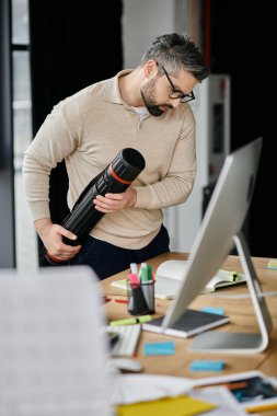 A bearded businessman wearing glasses stands in a modern office with a large computer screen, looking at a tube he is holding. clipart