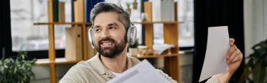 Sakallı yakışıklı bir işadamı modern bir ofiste çalışıyor, kulaklık takarken belgeleri inceliyor..