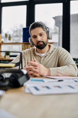 Sakallı yakışıklı bir işadamı modern bir ofis masasında oturuyor, kulaklık takıyor ve dikkatle belgelere bakıyor..