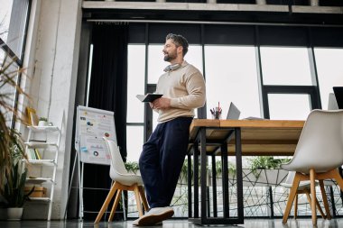 A handsome businessman with a beard stands in a modern office, holding a notebook, contemplating his next move. clipart