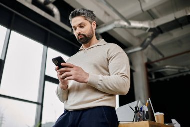 Sakallı yakışıklı bir işadamı modern bir ofiste dururken telefonunu kontrol ediyor..