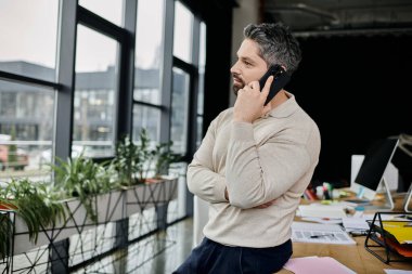 Yakışıklı sakallı bir işadamı modern bir ofiste duruyor, telefonuyla konuşuyor, kollarını kavuşturup pencereden dışarı bakıyor..