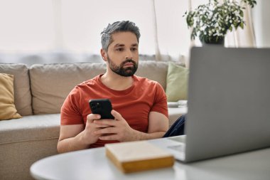 Sakallı bir adam dizüstü bilgisayarında çalışırken diğer elinde bir akıllı telefon tutuyor..