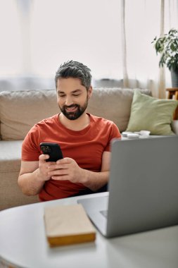 Günlük kıyafetli sakallı bir adam evinde uzaktan çalışıyor, akıllı telefonunu kullanıyor ve yanında bir dizüstü bilgisayar oturuyor..