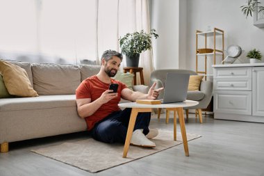 Sakallı bir adam oturma odasında yerde oturup dizüstü bilgisayarıyla çalışıyor. Elinde bir telefon tutuyor ve bilgisayarın ekranına bakıyor..