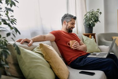 Sakallı bir adam evdeki koltukta rahatça oturur ve laptopuyla çalışır..