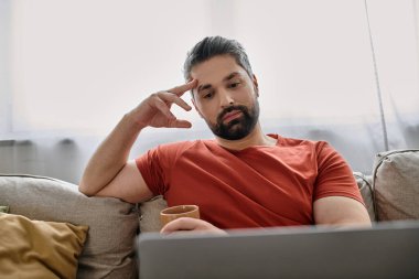 A bearded man in casual attire works remotely from home on a couch, using a laptop. clipart