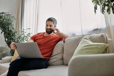 Günlük kıyafetli sakallı bir adam telefonuyla konuşurken bilgisayarla evden uzaktan çalışıyor..