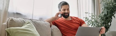 Günlük kıyafetli sakallı bir adam kanepede oturur, dizüstü bilgisayarla çalışır ve telefonuyla konuşur. Arkasındaki pencereden içeri doğal ışık giriyor..