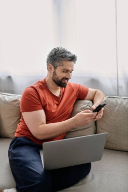 Sakallı bir adam evinde kanepede oturur, telefonunu kontrol ederken dizüstü bilgisayarda çalışır..