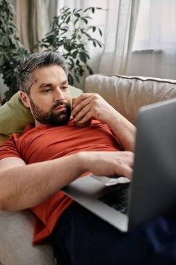 Günlük giysiler içinde sakallı bir adam evden uzakta, koltukta oturuyor ve dizüstü bilgisayar kullanıyor..