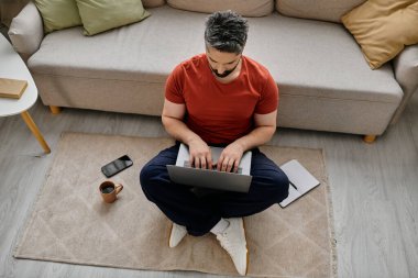 A man with a beard is sitting on the floor in front of a sofa, working on a laptop. He is wearing casual clothes and appears to be working from home. clipart