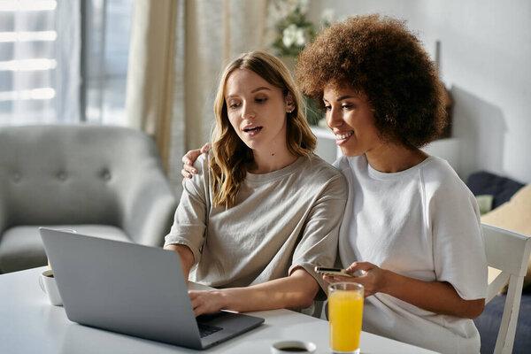 A lesbian couple relaxes at home, one working on a laptop while the other holds a phone.