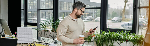 Красивый бизнесмен с бородой проверяет телефон, держа чашку кофе в современном офисе..