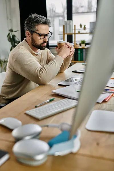 stock image A handsome businessman with a beard sits at his desk in a modern office, working on a project while enjoying a cup of coffee.