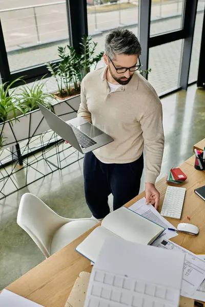 stock image A handsome bearded businessman in a modern office is holding a laptop and reviewing documents.