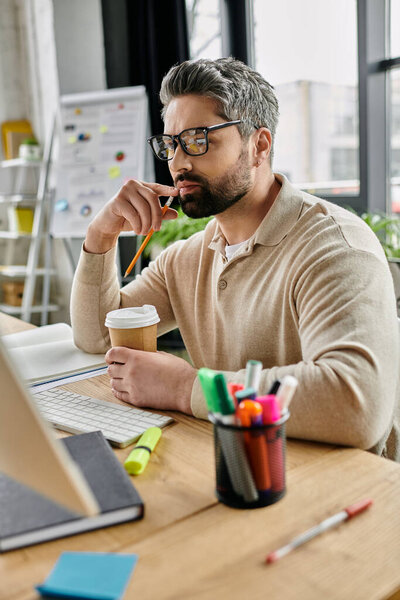 Красивый бизнесмен с бородой обдумывает проект, сидя за своим столом в современном офисе. Он держит чашку кофе в одной руке и карандаш в другой..