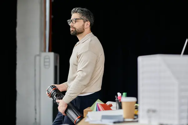 stock image A handsome businessman with a beard stands in a modern office, holding a blueprint and thoughtfully gazing away.