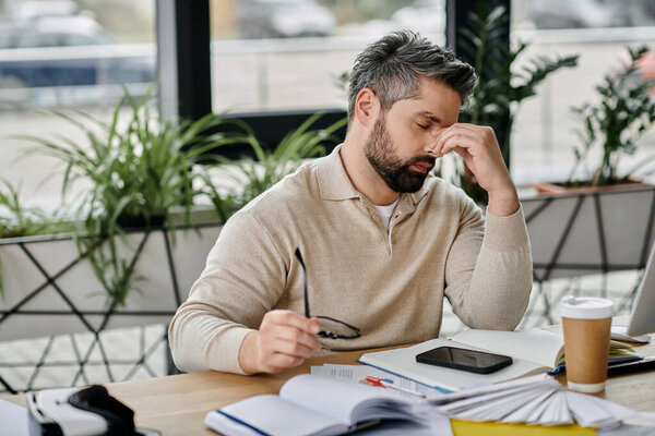 Бородатый бизнесмен в современном офисе, казалось бы потерявшийся в мыслях, сидя за своим столом, окруженный бумажной работой, смартфоном и чашкой кофе.