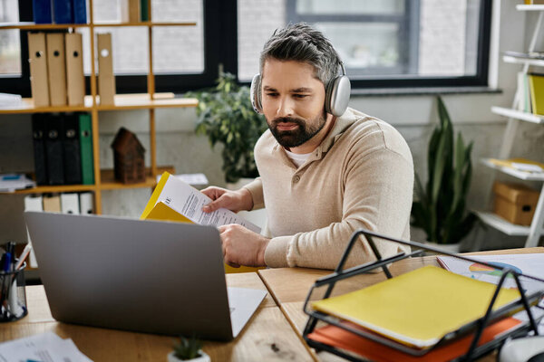 Бизнесмен с бородой сидит за своим столом в современном офисе, просматривая документы в наушниках.