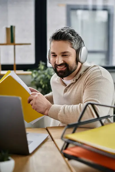 stock image A businessman with a beard works diligently in a modern office, with a laptop, files, and headphones.