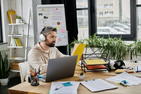 Kulaklık takan sakallı bir iş adamı, modern bir ofiste masasında çalışıyor. Bilgisayarına ve sarı dosyasına odaklanmış durumda..