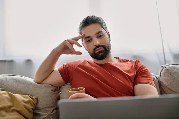 stock image A bearded man in casual attire works remotely from home on a couch, using a laptop.