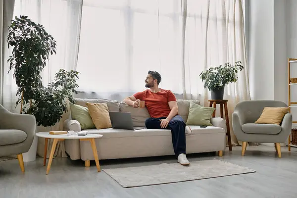stock image A bearded man in casual attire works remotely from his home office, sitting on a couch with a laptop and a cup of coffee.