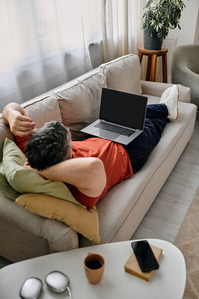 A bearded man in casual attire lies on a white couch with his feet up, working remotely on his laptop.