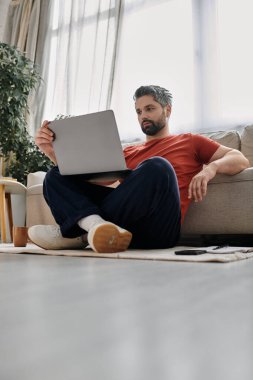 Günlük giysiler içinde sakallı bir adam elinde dizüstü bilgisayarla yerde oturuyor ve uzaktan çalışıyor..