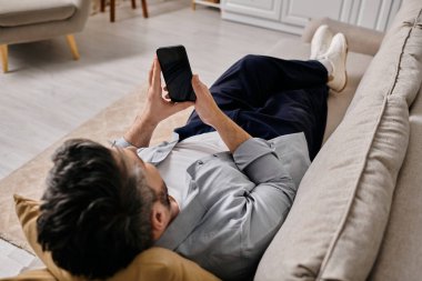 Günlük giysiler içinde sakallı bir adam kanepeye uzanıp akıllı telefonunu karıştırıyor..