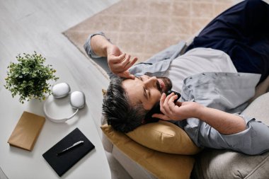 Günlük giysiler içinde sakallı bir adam kanepede uzanıyor, evde dinlenirken telefonla konuşuyor..