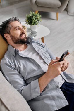 Gri tişörtlü sakallı bir adam evinden kanepesinde çalışıyor, elinde akıllı bir telefon var..