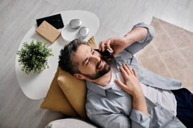 Düğmeli gömlekli sakallı bir adam evdeki bir koltukta oturmuş, telefonla gülüyor..