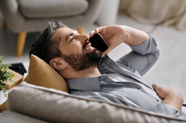 Sıradan giyinmiş sakallı bir adam uzaktan çalışırken kanepeye uzanıp telefonuyla konuşuyor..