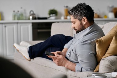 Sıradan giyinmiş sakallı bir adam kanepeye oturur, ayaklarını uzatıp telefonunu kontrol eder..