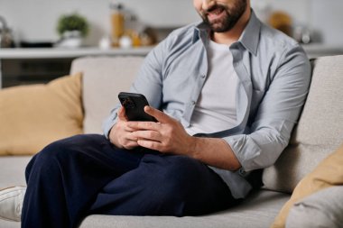 A bearded man wearing casual clothing is sitting on a couch, working remotely from home. He is using his smartphone. clipart