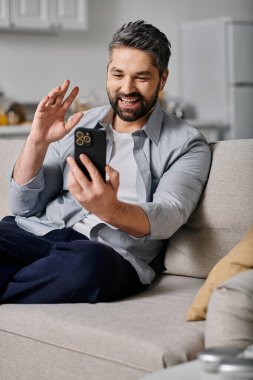 Günlük kıyafetli sakallı bir adam kanepede oturur ve video sohbeti yaparken telefonuna el sallar..